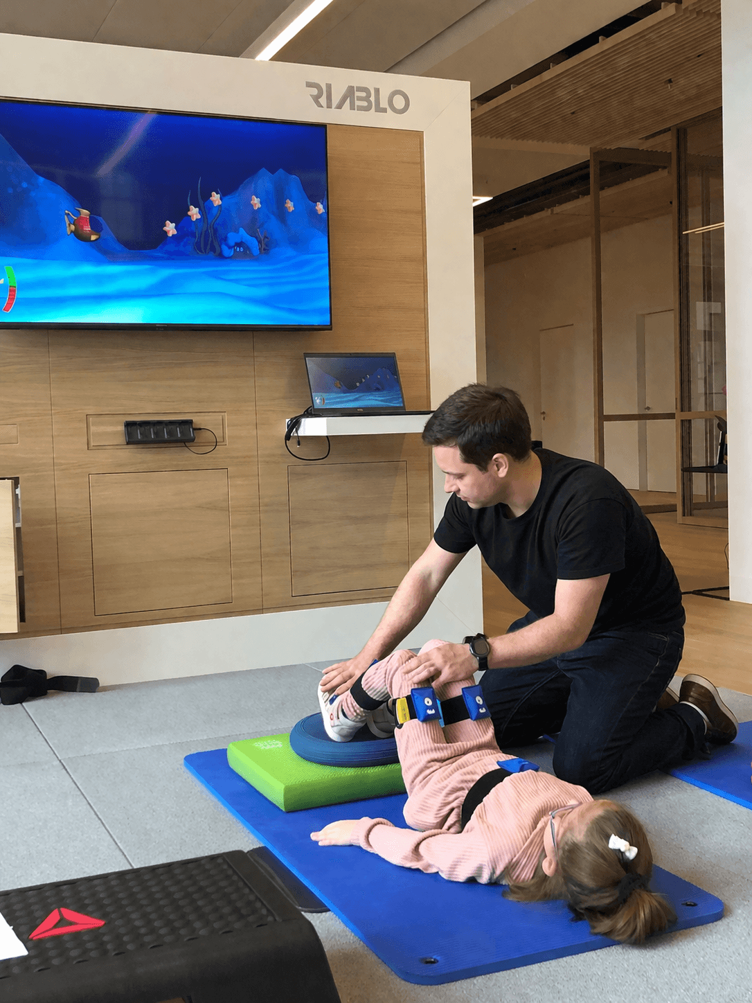 A child lies on a mat with Riablo sensors on her legs while a therapist guides the exercise